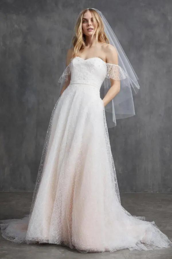 This romantic blush off-the-shoulder bridal gown features approximately 1600 pearls and takes our team of talented seamstresses over 30 hours to hand bead. You’ll love the spider web detail and lattice work on Ruby, plus the pockets make this flutter sleeve wedding dress a no brainer | Marilyn Collection by Kelly Faetanini
